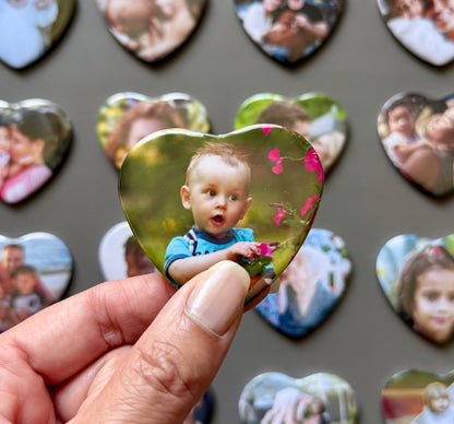Heart-shaped photo magnet of a child held by a hand with more magnets in the background.