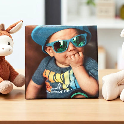 Square photo magnet of a child wearing sunglasses and a blue cap on a wooden surface with toys around.