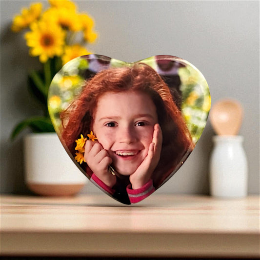 Heart-shaped photo magnet with a smiling girl's face on a wooden surface with flowers in the background
